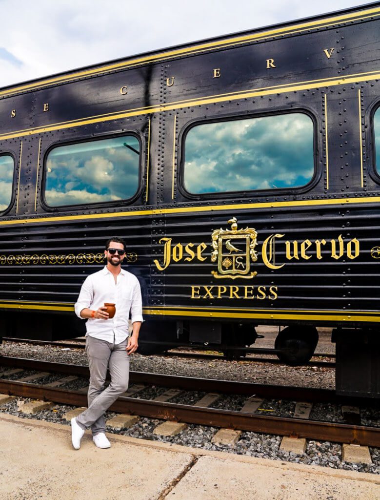 Man holding a drink and leaning against a train entitled the Jose Cuervo Express in the town of Tequila, Mexico
